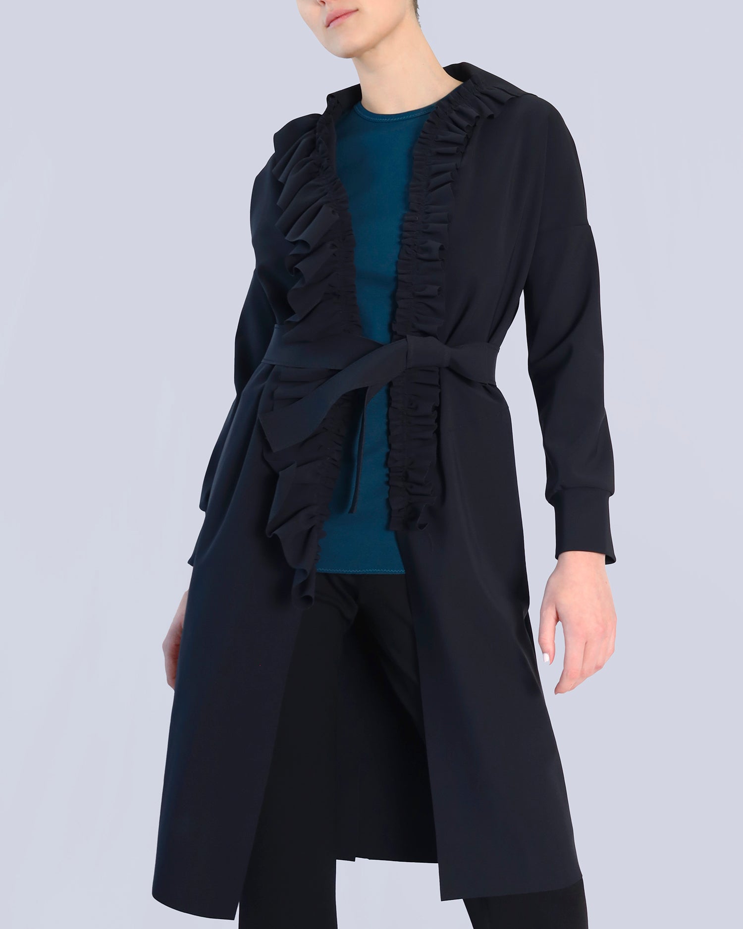 Person wearing a black long coat with ruffled details on a light gray background. Maison Marie Saint Pierre | Coat | PELLINAME | ANT | Pants | PALLAS | Black | Top | PALRAIR | Petrol